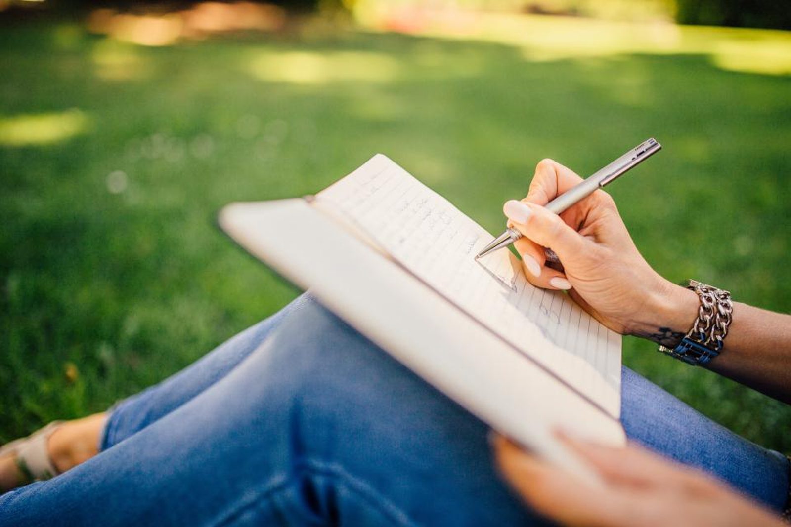 Écriture libératrice en plein air : une main tient un stylo au-dessus d'un carnet posé sur les genoux, dans l'herbe lumineuse